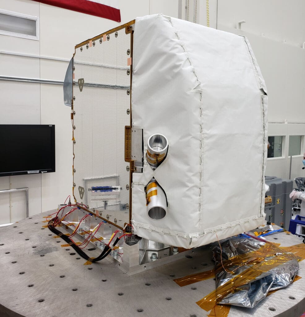 An imaging spectrometer stands waiting to be transferred in the NASA JPL lab. It has a white thermal cover over it. It's arrival is noted in the text around it.
